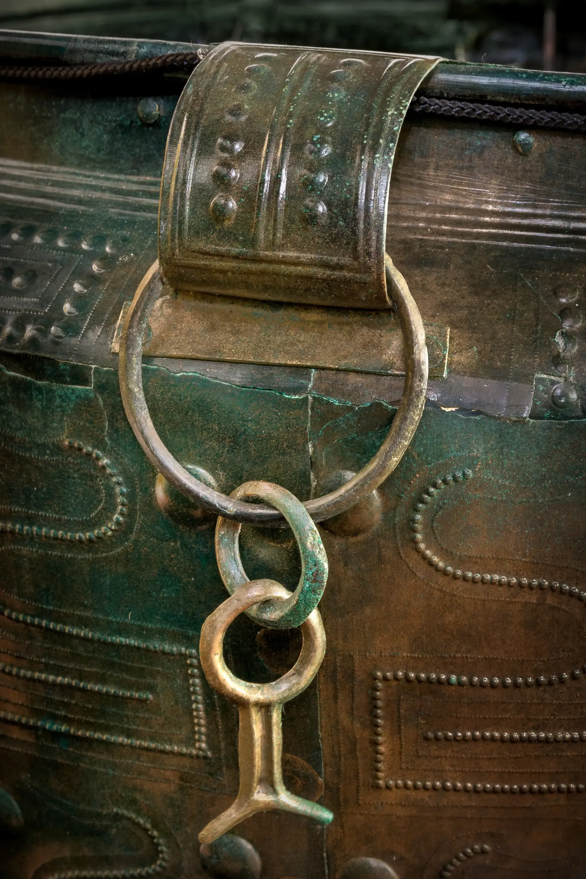 Detailaufnahme: In einem bandförmigen Henkel hängt ein Anhänger aus zwei Ringen und einem schwalbenschwanzförmigen Objekt. Alles aus grüner Bronze.