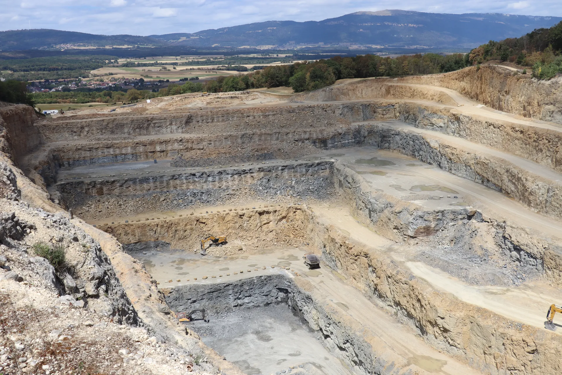 Ein großer Steinbruch mit mehreren, terrassenförmigen Abbaustufen, in dem vier Baumaschinen arbeiten. Im Hintergrund Felder und bewaldete Berge.