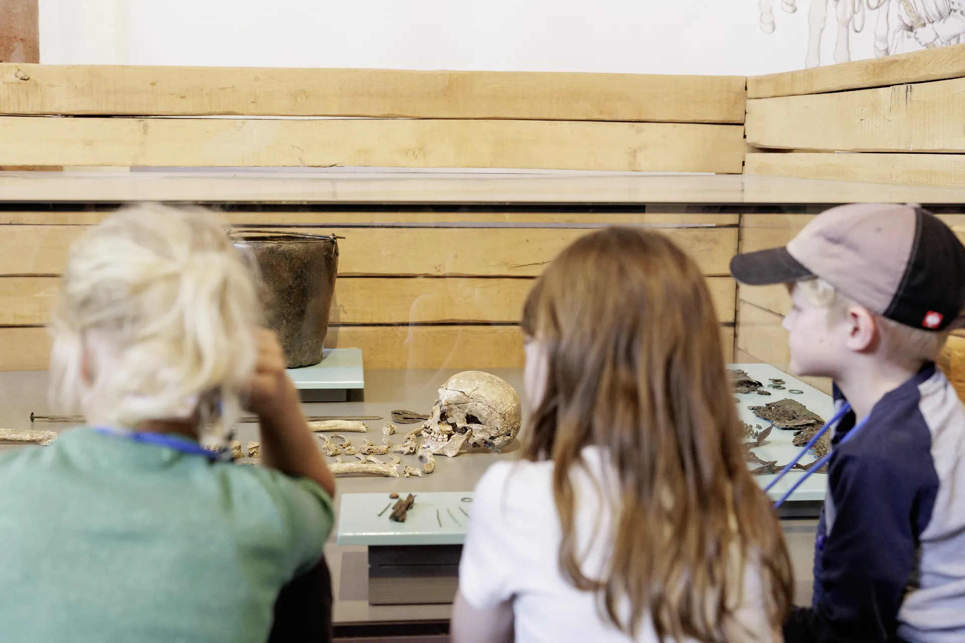 Drei Kinder betrachten bei einer Führung eine Bestattung mit Beigaben in einer Vitrine aus Glas und Holz.