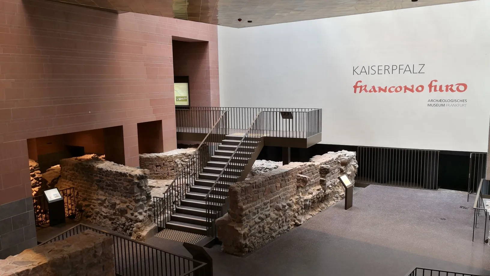 Blick in einen modernen Ausstellungsraum mit vielen alten Mauerresten von Gebäuden aus Stein, Ausstellungstafeln sowie Geländern und Treppen. An der Wand ein Schriftzug: Kaiserpfalz Franconofurd Archäologisches Museum Frankfurt.