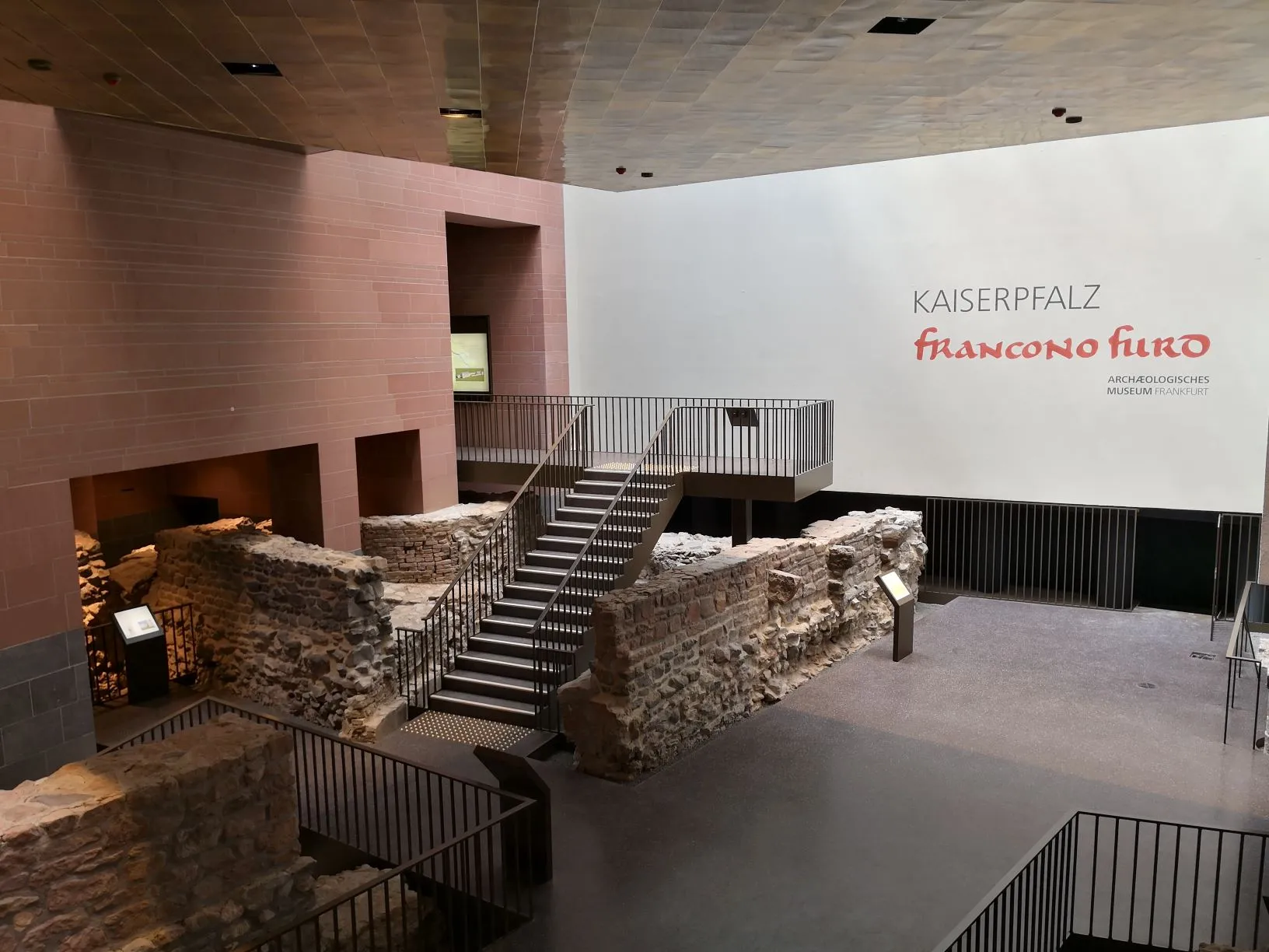 Blick in einen modernen Ausstellungsraum mit vielen alten Mauerresten von Gebäuden aus Stein, Ausstellungstafeln sowie Geländern und Treppen. An der Wand ein Schriftzug: Kaiserpfalz Franconofurd Archäologisches Museum Frankfurt.
