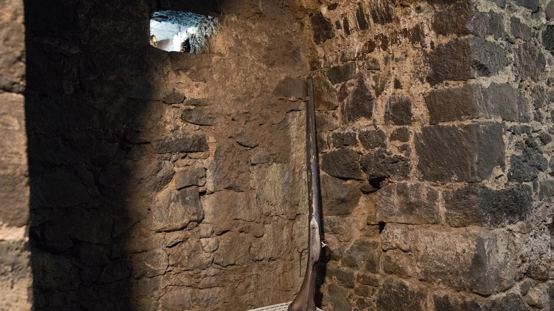 Ein altes Gewehr lehnt in einer steingemauerten Nische an der Wand. Sie wird durch ein kleines Fenster erhellt.