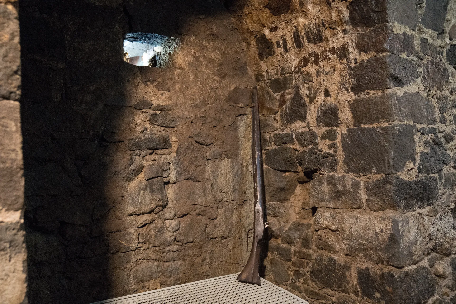 Ein altes Gewehr lehnt in einer steingemauerten Nische an der Wand. Sie wird durch ein kleines Fenster erhellt.