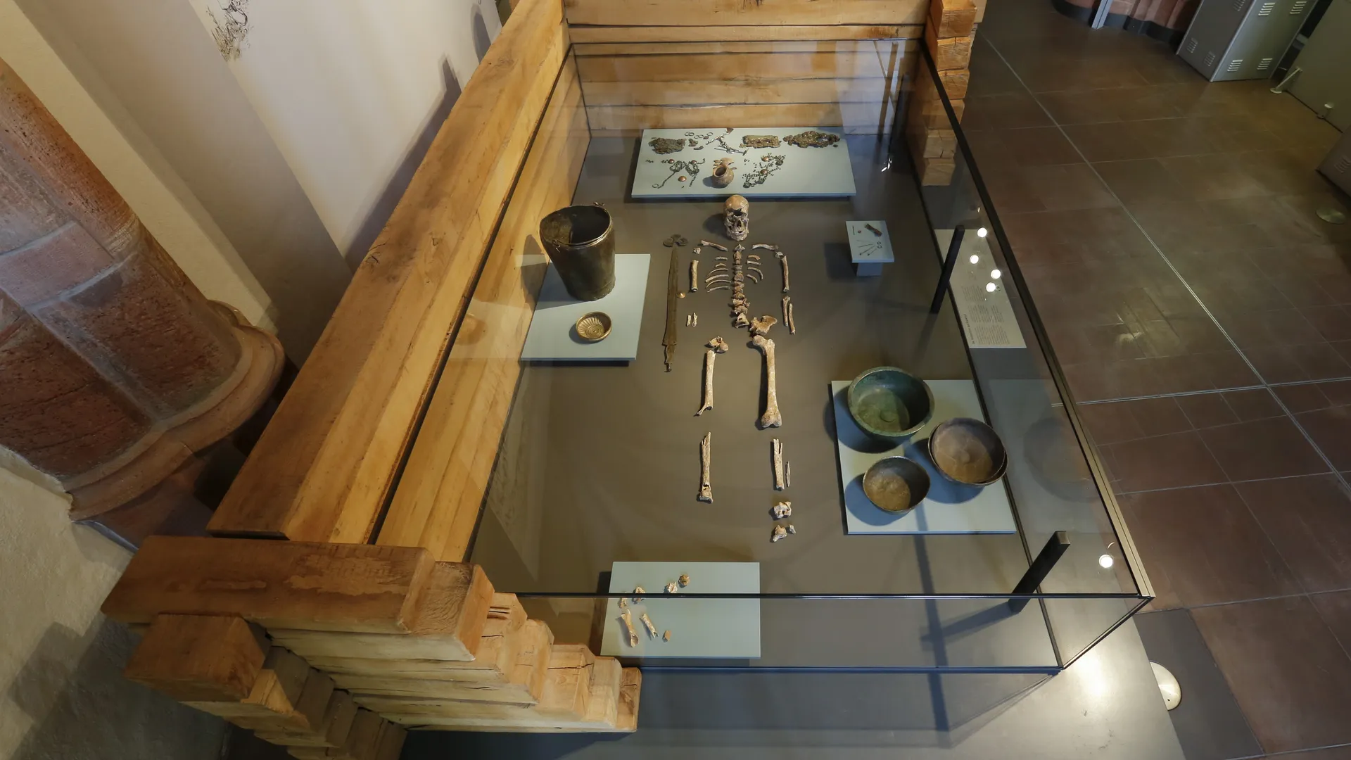 Blick von oben in eine Vitrine aus Holz und Glas in einem Ausstellungsraum. Sie enthält ein menschliches Skelett in der Mitte. Darum herum liegen verschiedene Grabbeigaben wie Gefäße, Knochen und Metallobjekte.