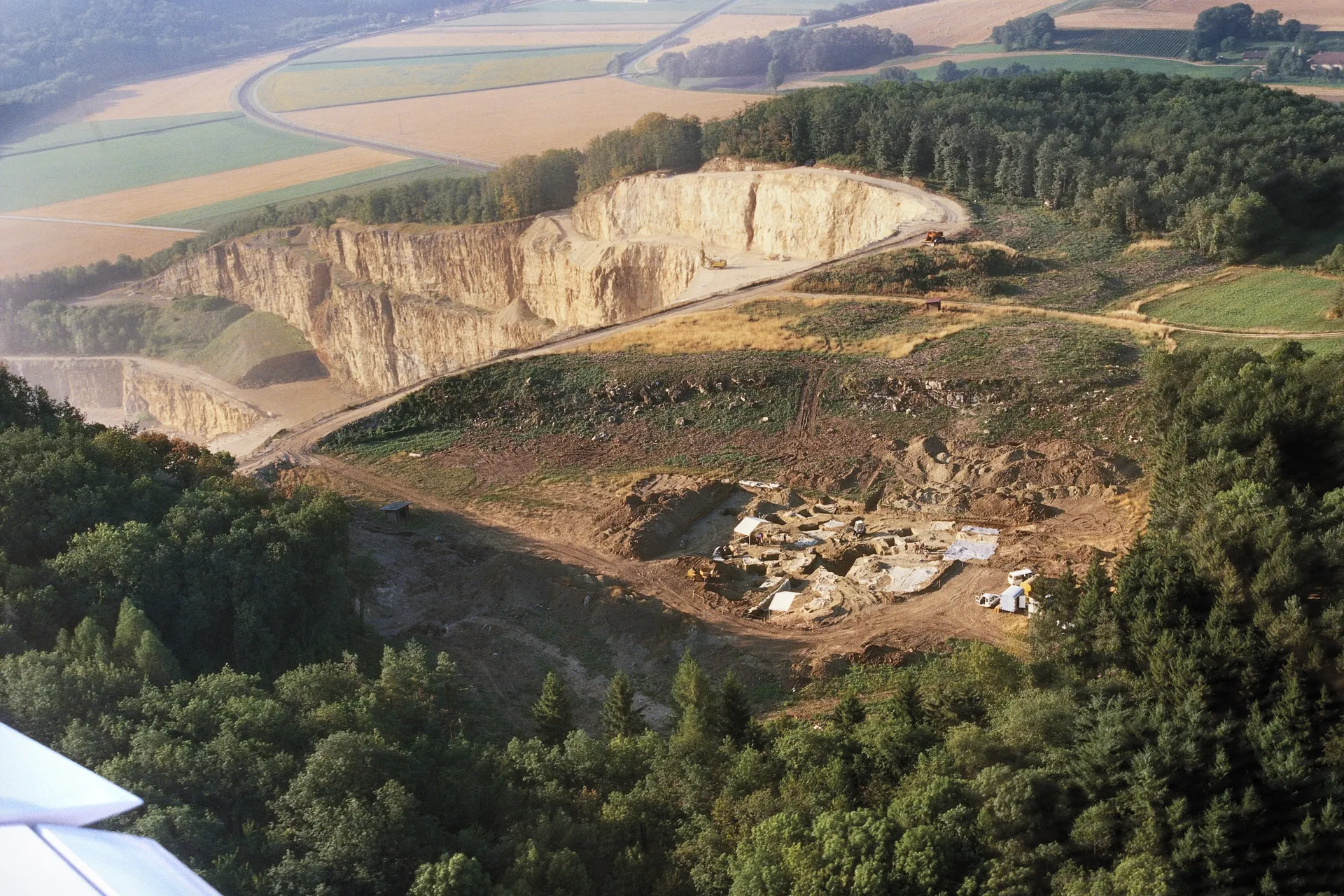 Eine archäologische Ausgrabungsfläche mit Zelten, Fahrzeugen und Grabungsgerät. Dahinter liegt ein großer Steinbruch. Im Hintergrund Bäume und Felder.