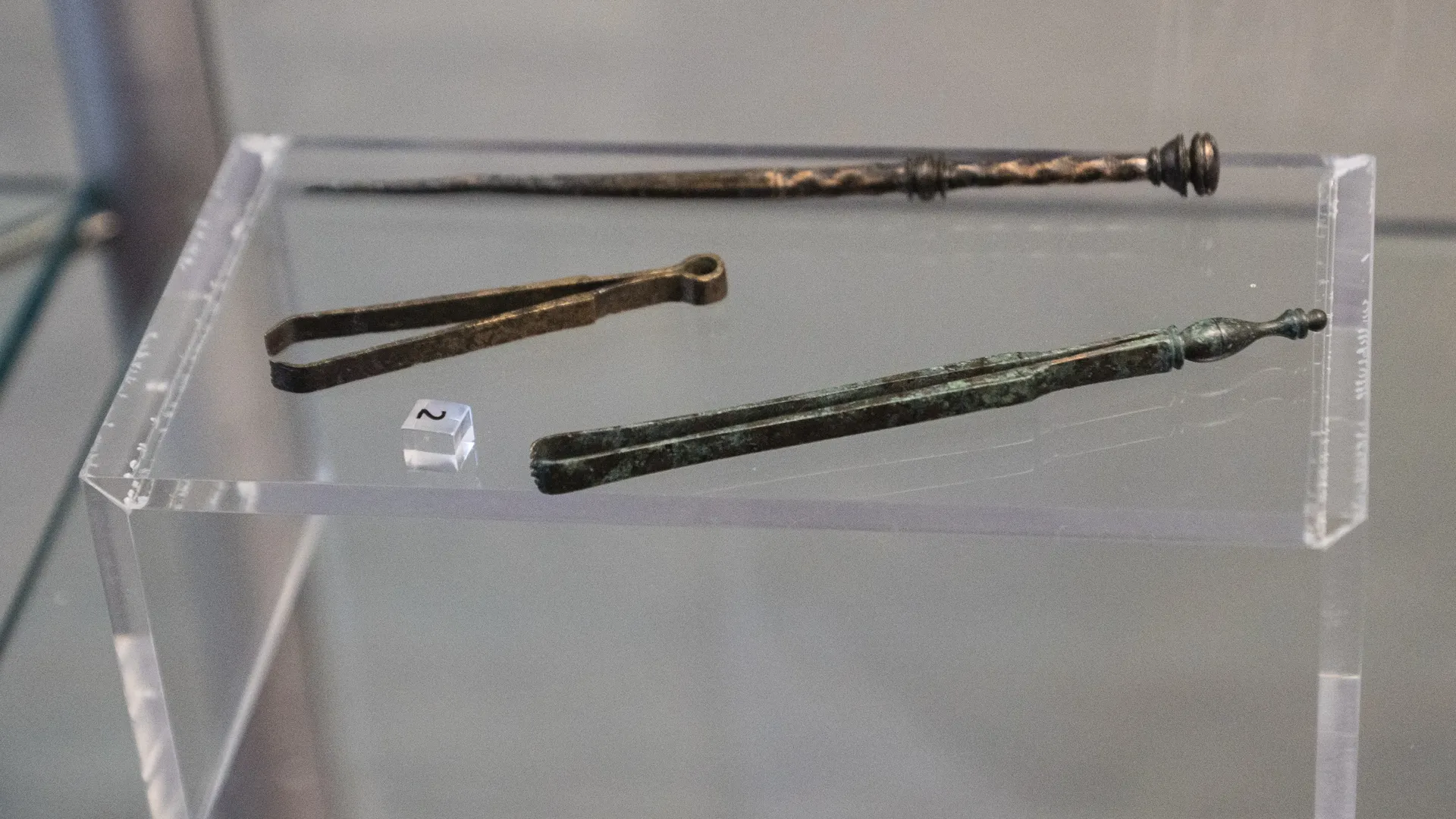 Zwei unterschiedlich große Pinzetten und ein spitzer Stab aus Bronze liegen nebeneinander in einer Vitrine.