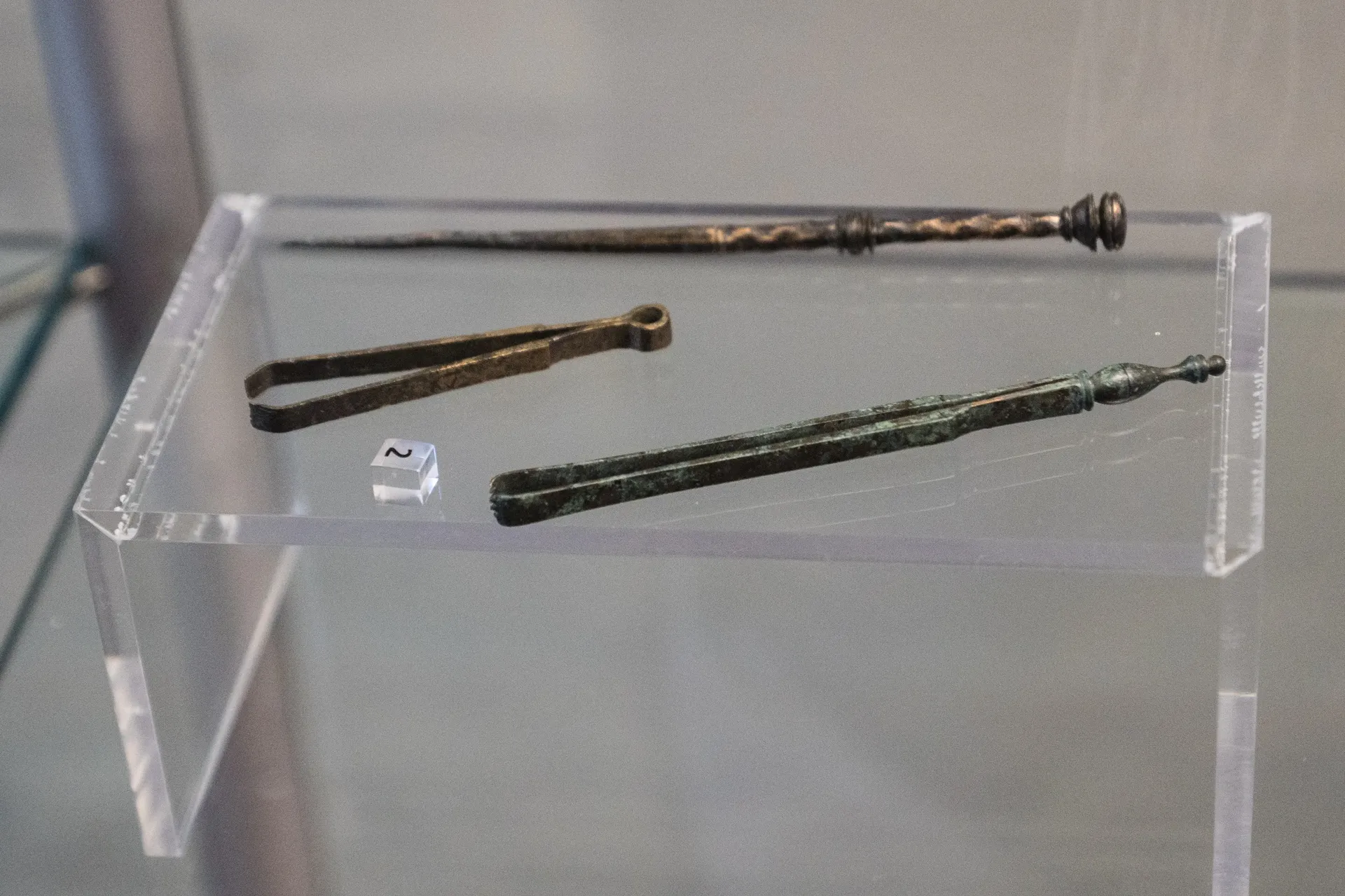 Zwei unterschiedlich große Pinzetten und ein spitzer Stab aus Bronze liegen nebeneinander in einer Vitrine.